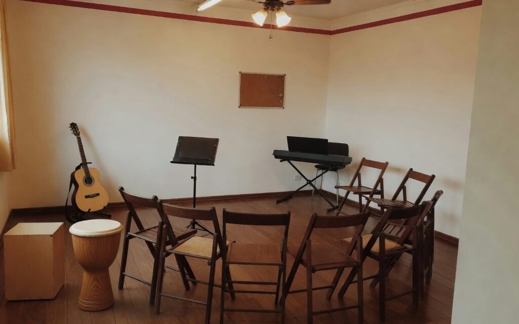 Sala de ensaio da EArte preparada para grupo, com cadeiras em semicírculo, estante de partitura, cajón, djembê, violão e teclado